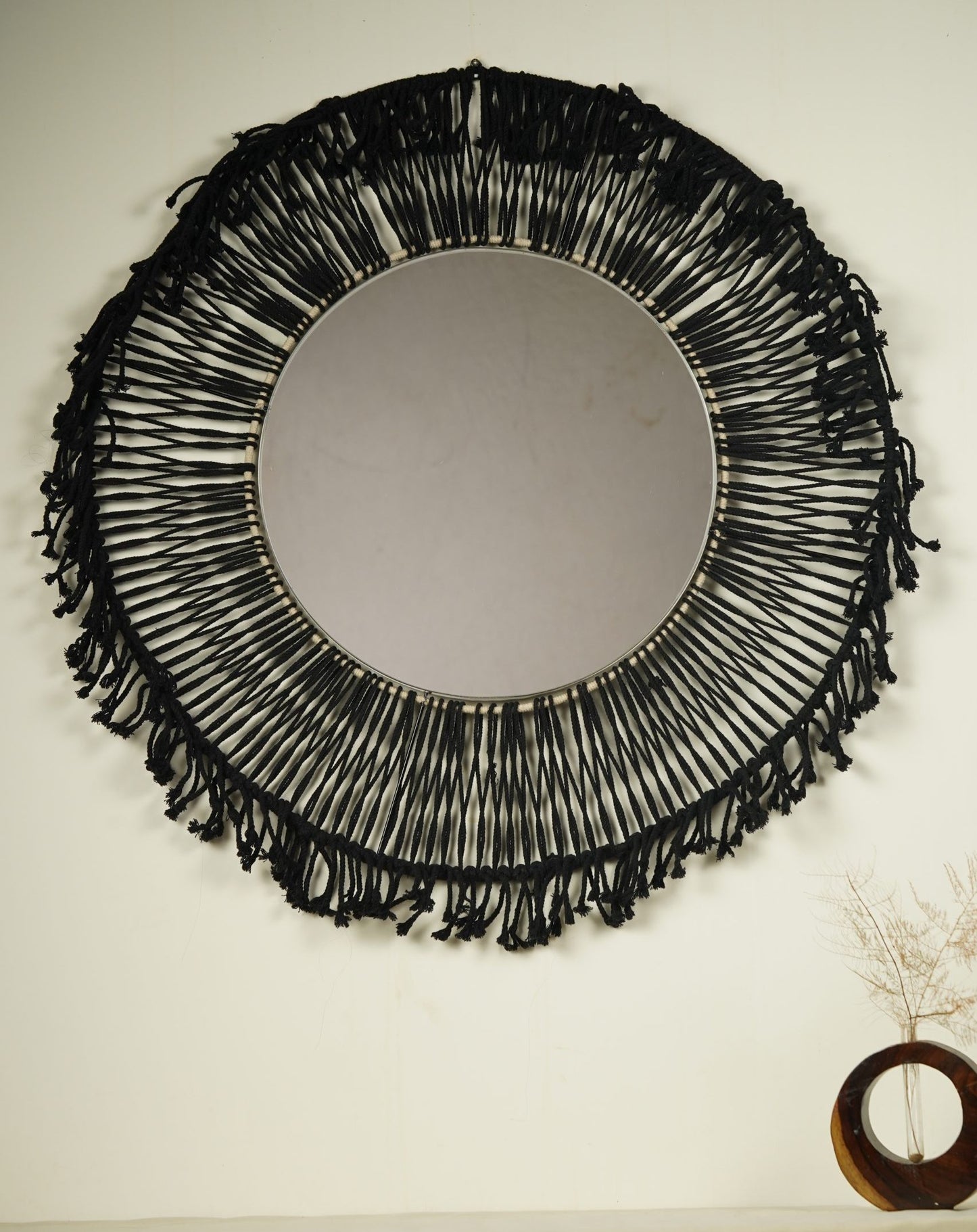 A round, black fringed hanging mirror with a visible pattern of crochet yarns.