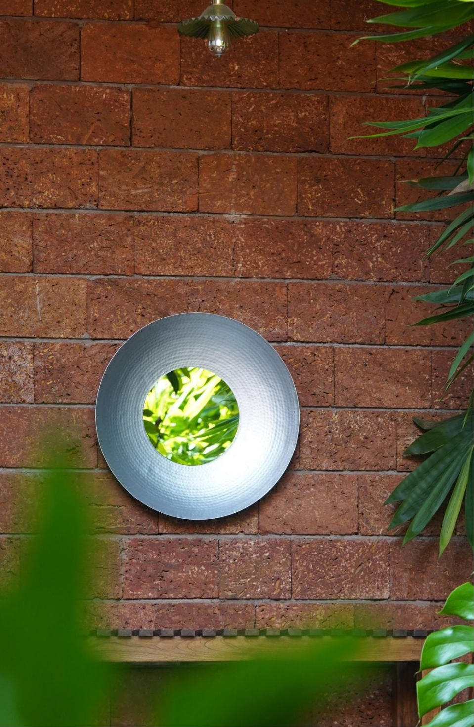 A round, silver hammered wall mirror hanging on a wall above a wooden table with a teapot and a plant.