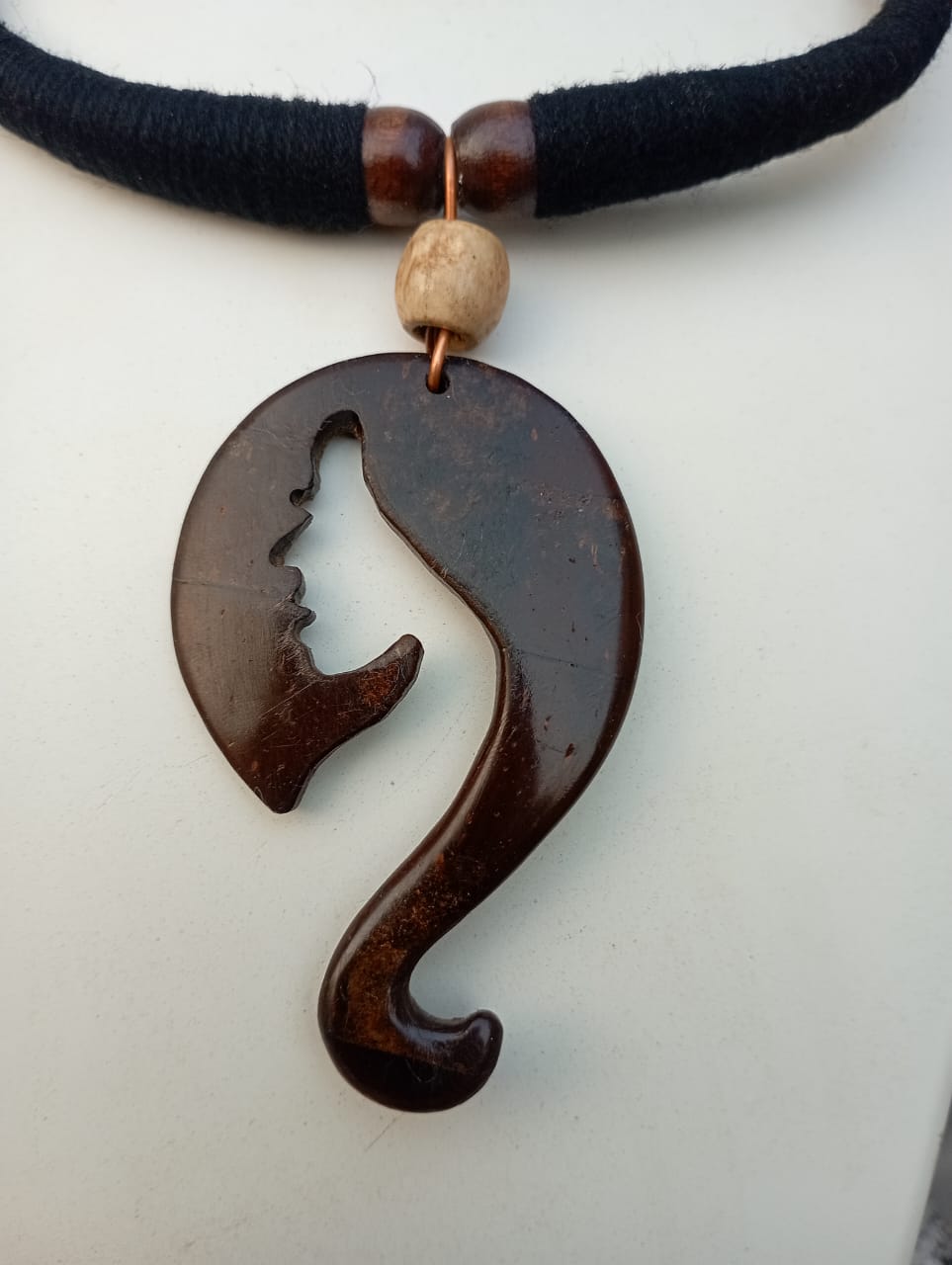A brown pendant necklace with a figure of a question mark on a coconut shell, displayed on a mannequin bust with a tropical background.
