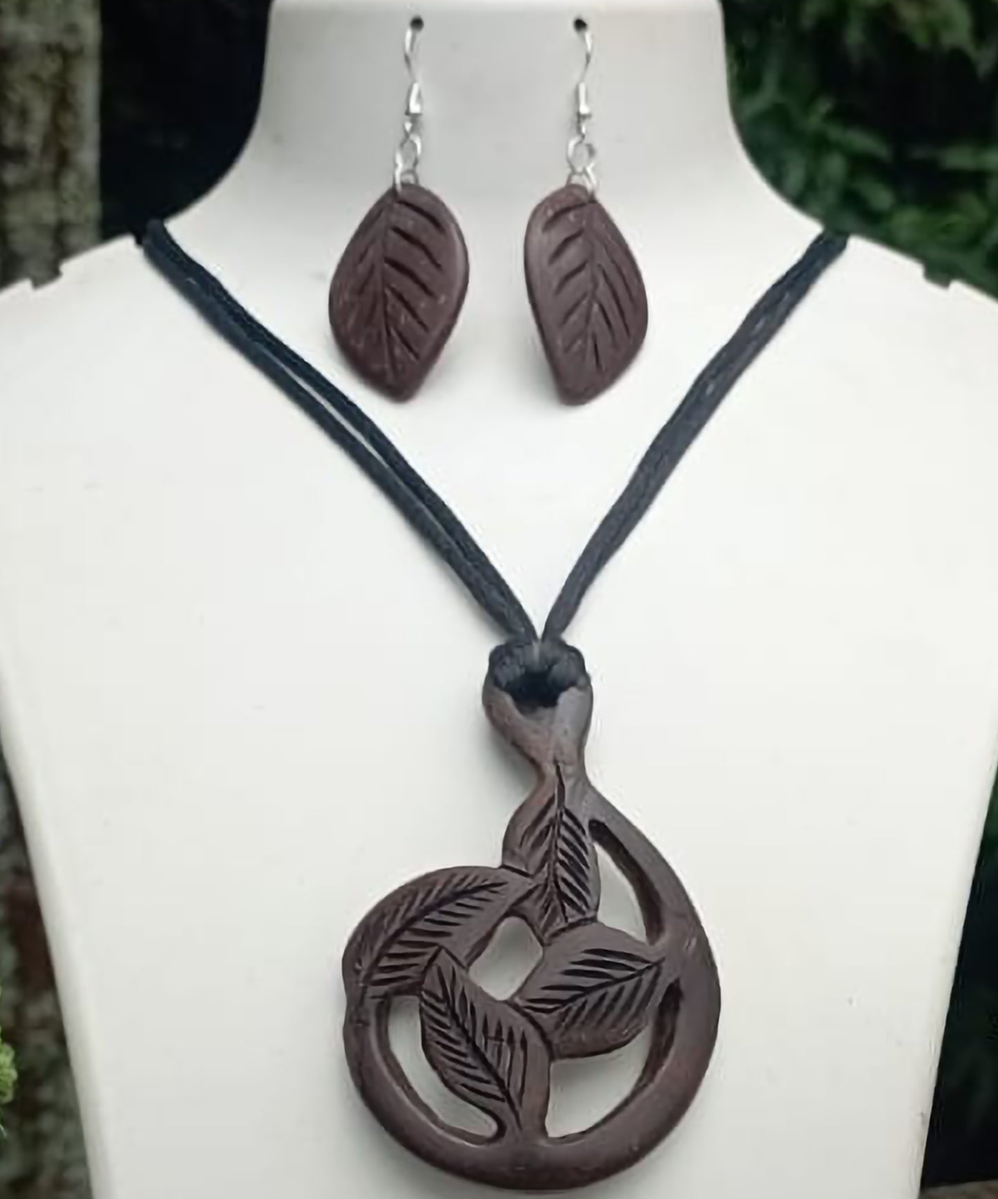 A pendant set with one pendant displayed on a mannequin bust and two coconut shell earrings on either side, set against a backdrop of plants.