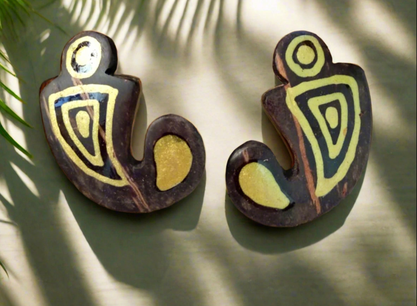 A pair of brown coconut shell earrings with gold geometric patterns, displayed against a wooden background with a plant in the background.
