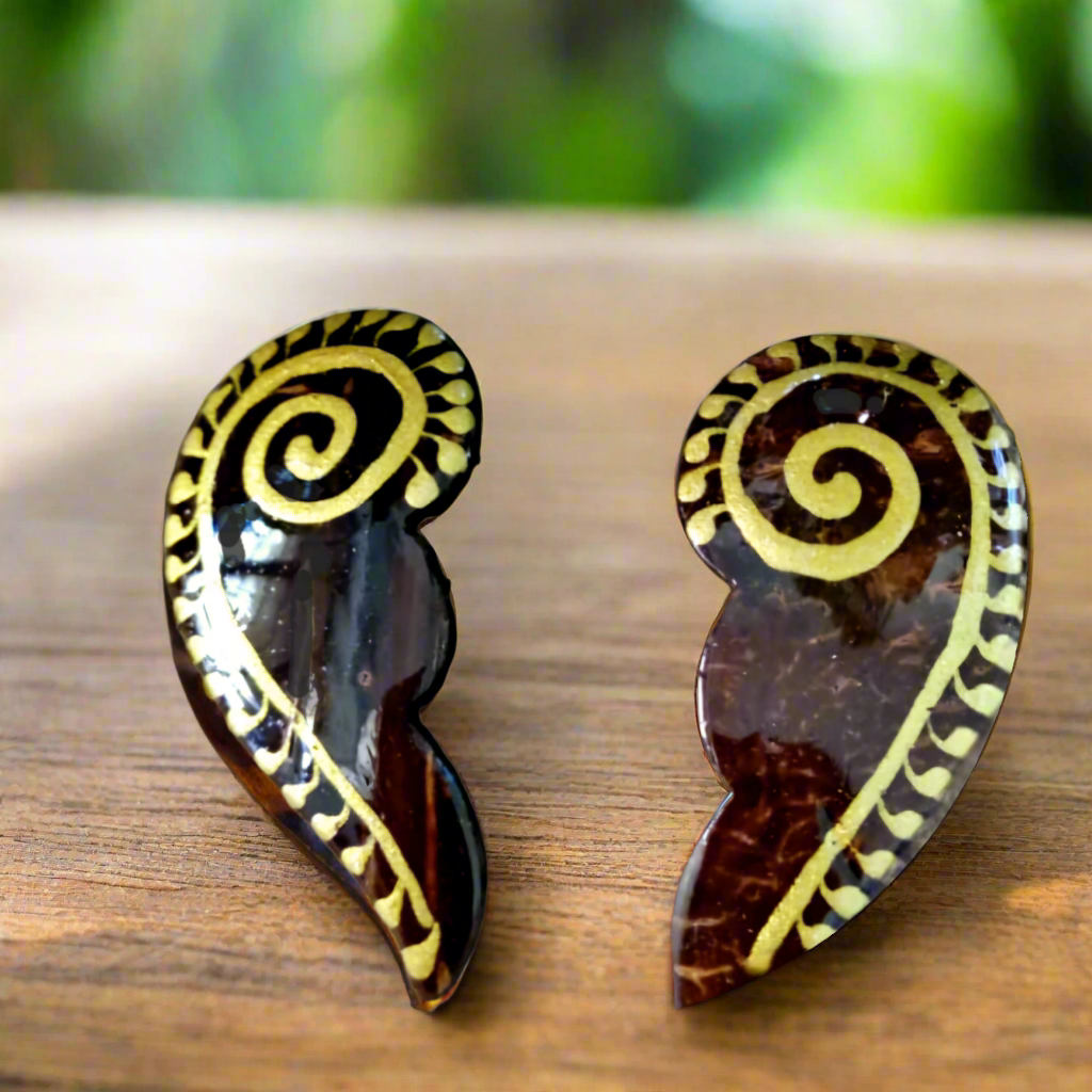 A pair of brown coconut shell earrings with gold accents on a wooden surface, with green leaves in the background.