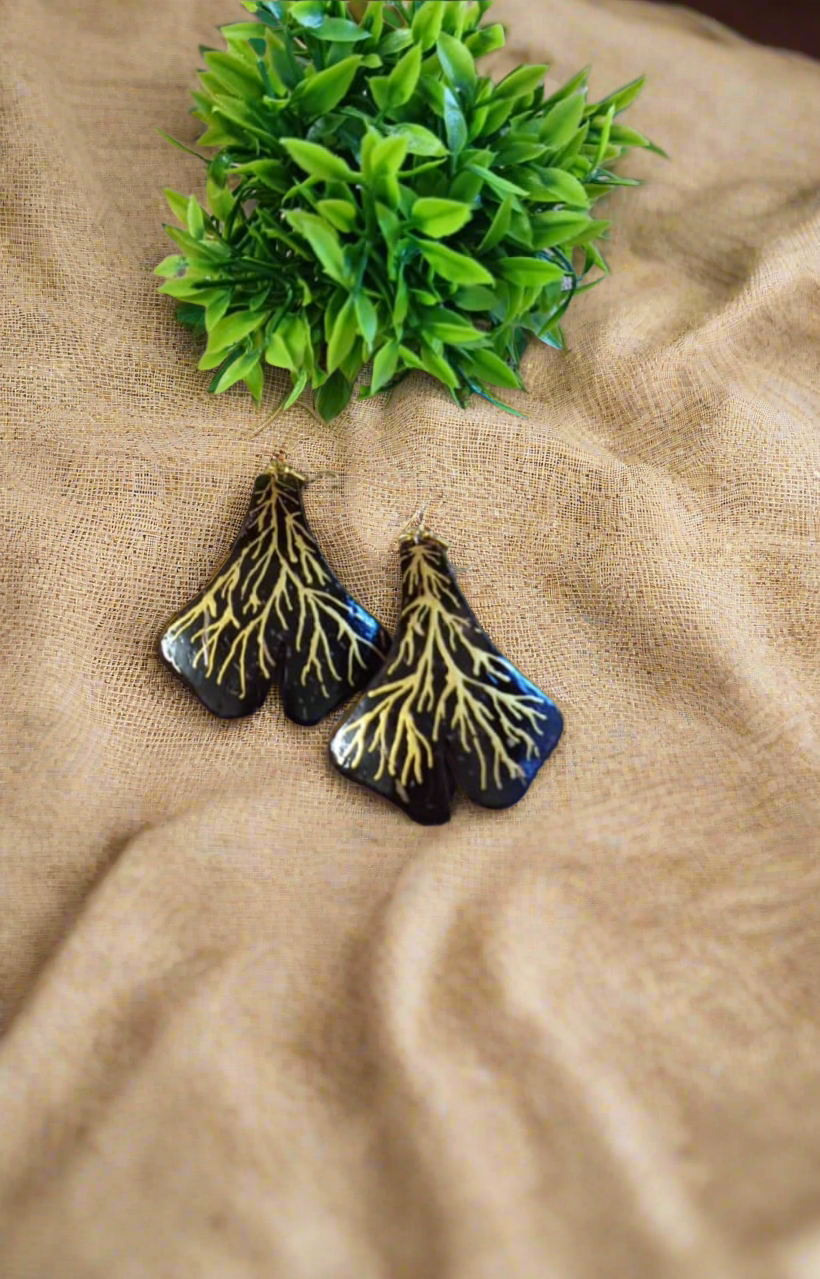 A pair of earrings designed to resemble tree roots, placed on a beige fabric surface with a green leafy object to the side.