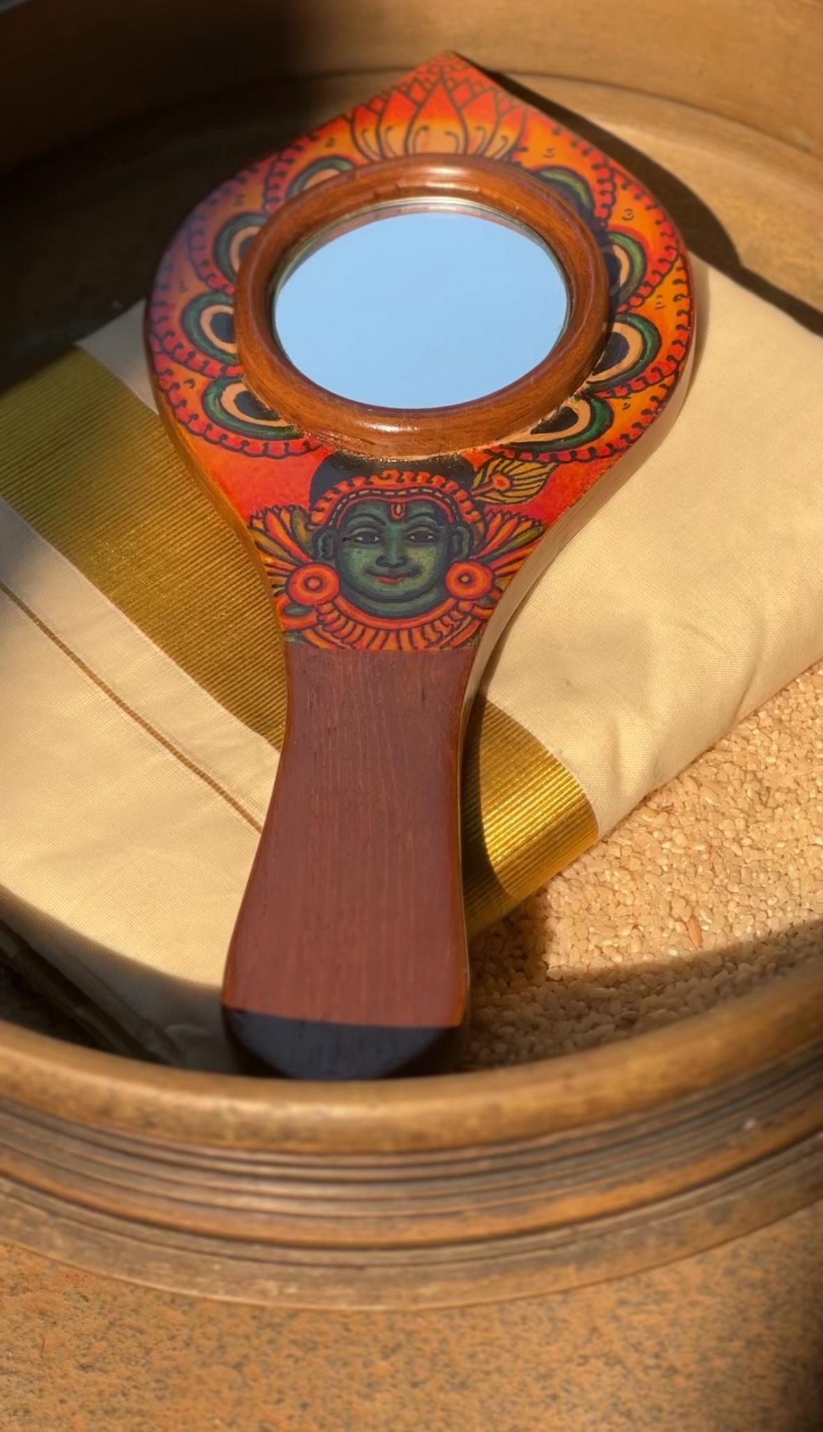 Handheld mirror with a colorful wooden frame featuring a traditional Kerala mural art depiction of Lord Krishna
