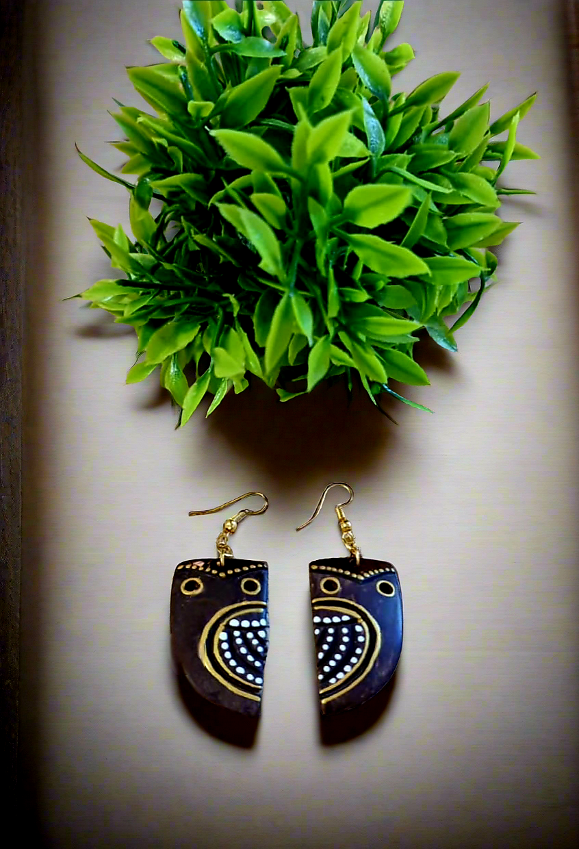 A pair of handpainted coconut shell earrings with a design resembling an owl's face, primarily black with white and gold details.