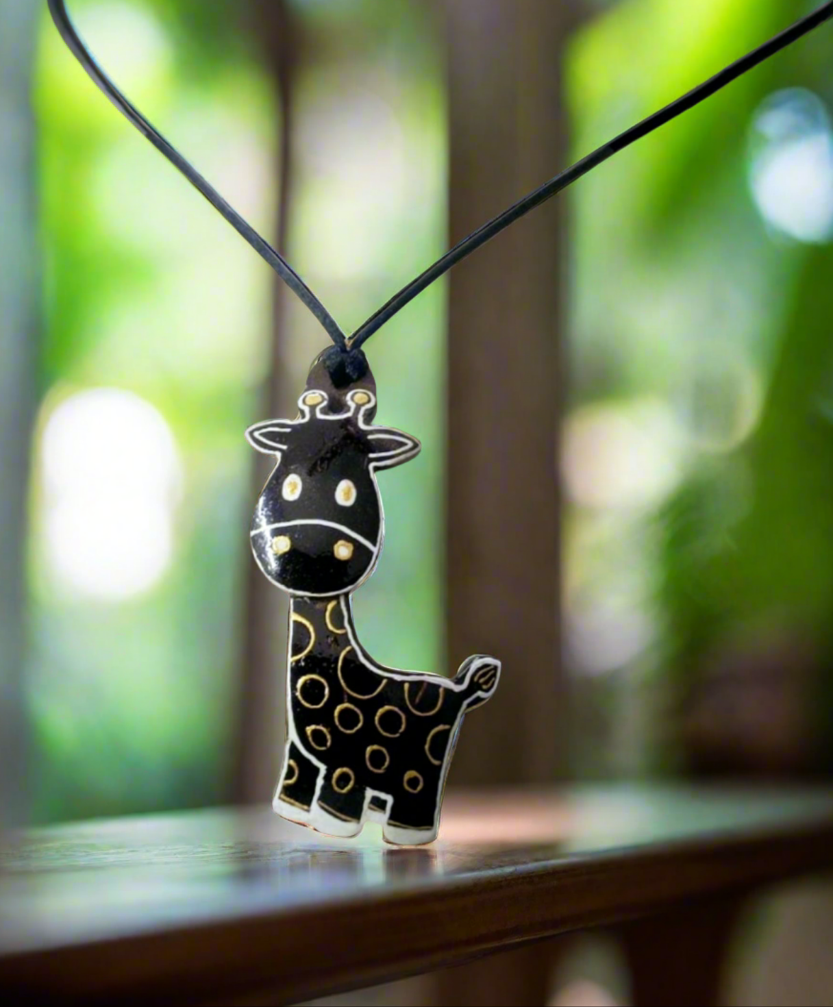 A necklace with a pendant in the shape of a giraffe with a black and gold pattern on a wooden surface with green foliage in the background.