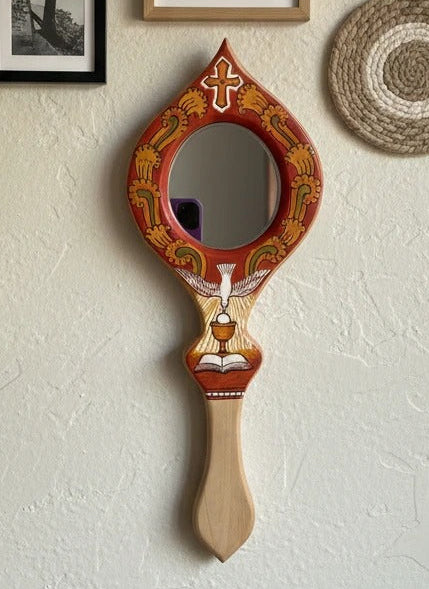 Decorative hand mirror with ornate frame on a wall with framed pictures and a woven basket.