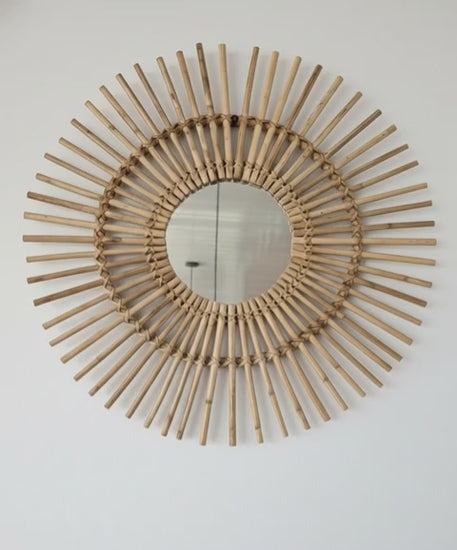 Decorative wall mirror with a sunburst design made of wooden sticks on a white wall.