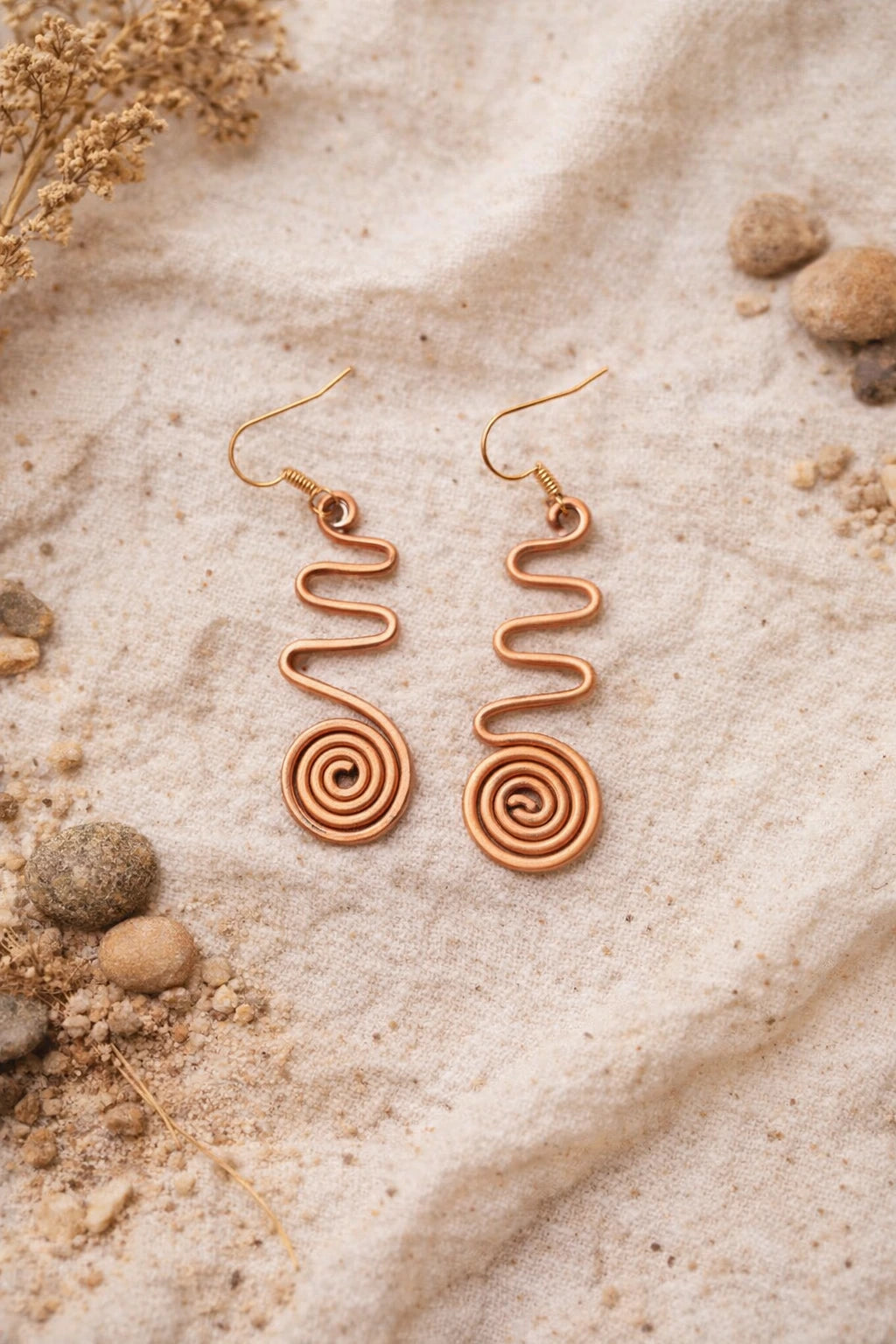 Rose gold spiral earrings on a sandy surface with dried flowers and rocks.