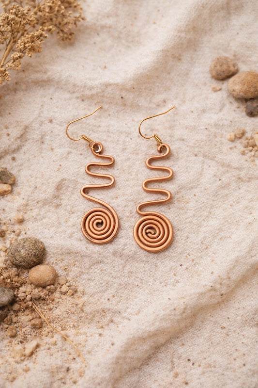 Rose gold spiral earrings on a sandy surface with dried flowers and rocks.