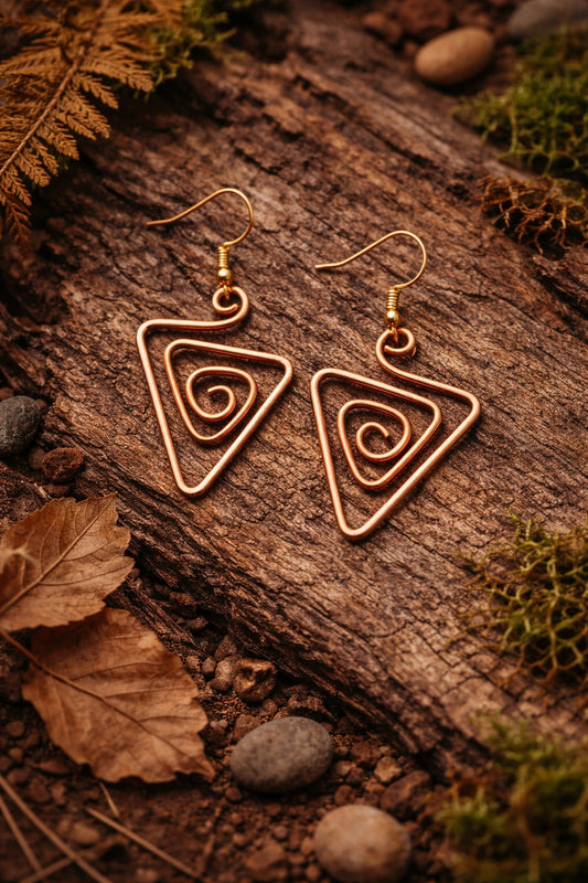Pair of gold spiral triangle earrings on a wooden surface with leaves and stones.