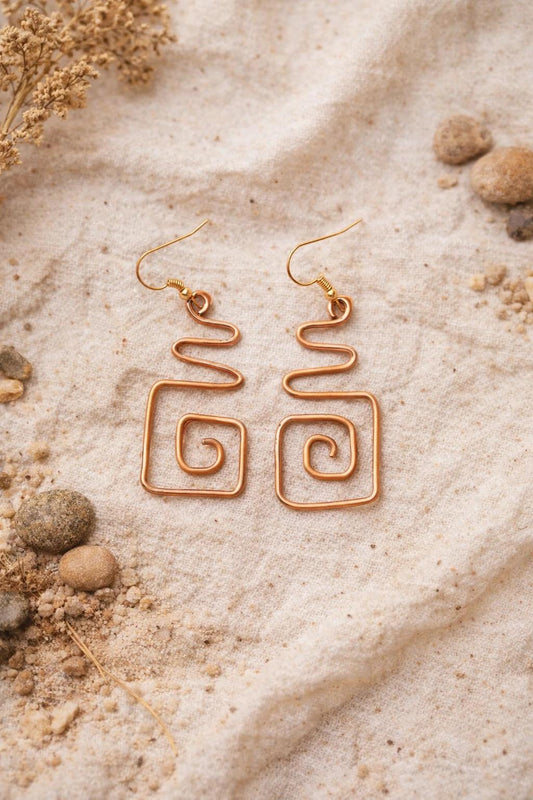 Gold spiral earrings on a sandy surface with dried plants.
